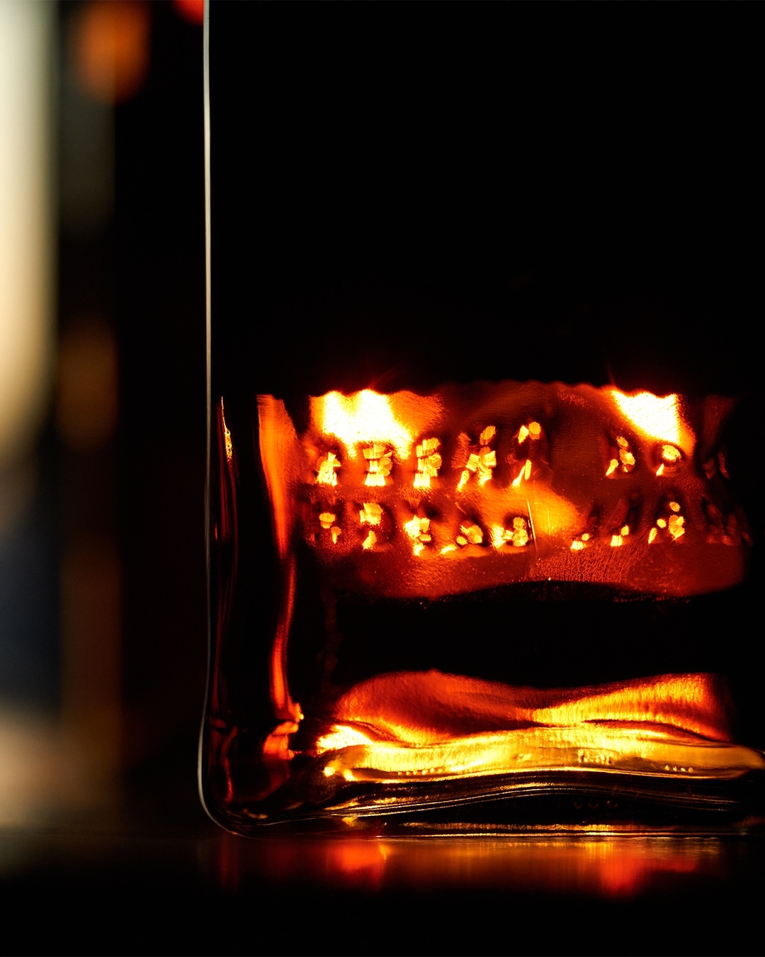 Close-up of an amber liquid-filled glass bottle on a dark surface, with light shining through the text embossed on its side.