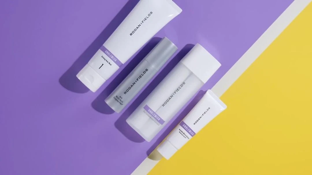 Rodan + Fields skincare products including a face wash, serum, and creams, arranged diagonally on a split purple and yellow background.