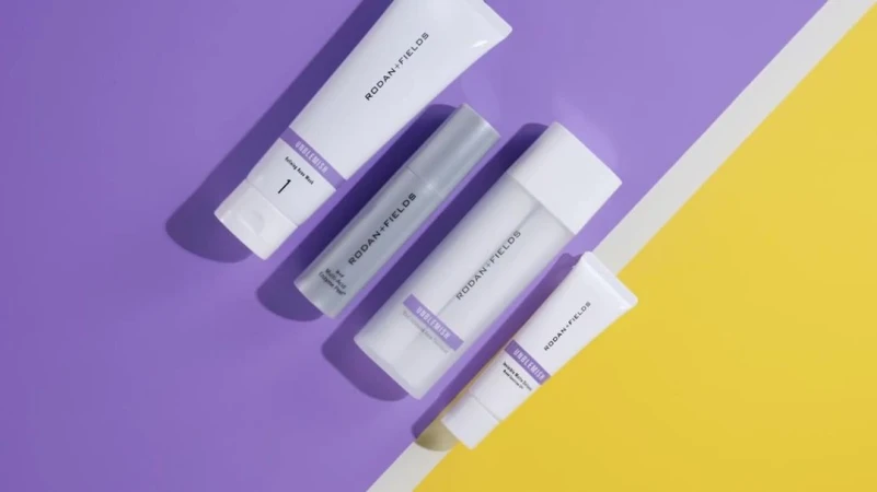 Rodan + Fields skincare products including a face wash, serum, and creams, arranged diagonally on a split purple and yellow background.