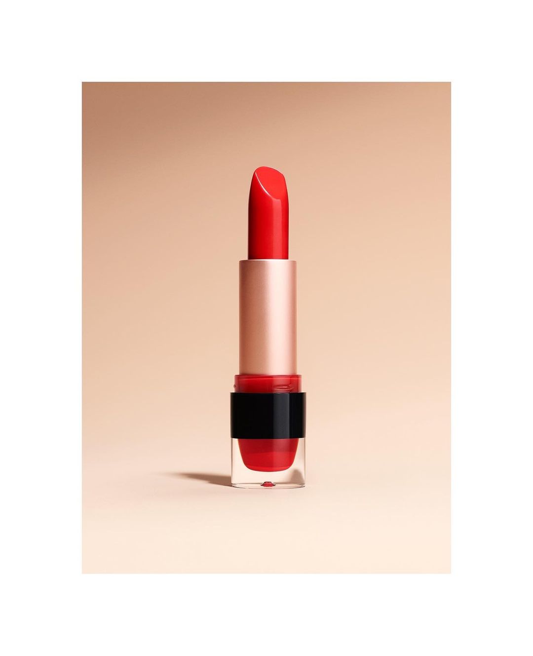 Close-up studio shot of a vibrant red lipstick with a gold tube and transparent base against a soft beige background. Beauty and makeup.