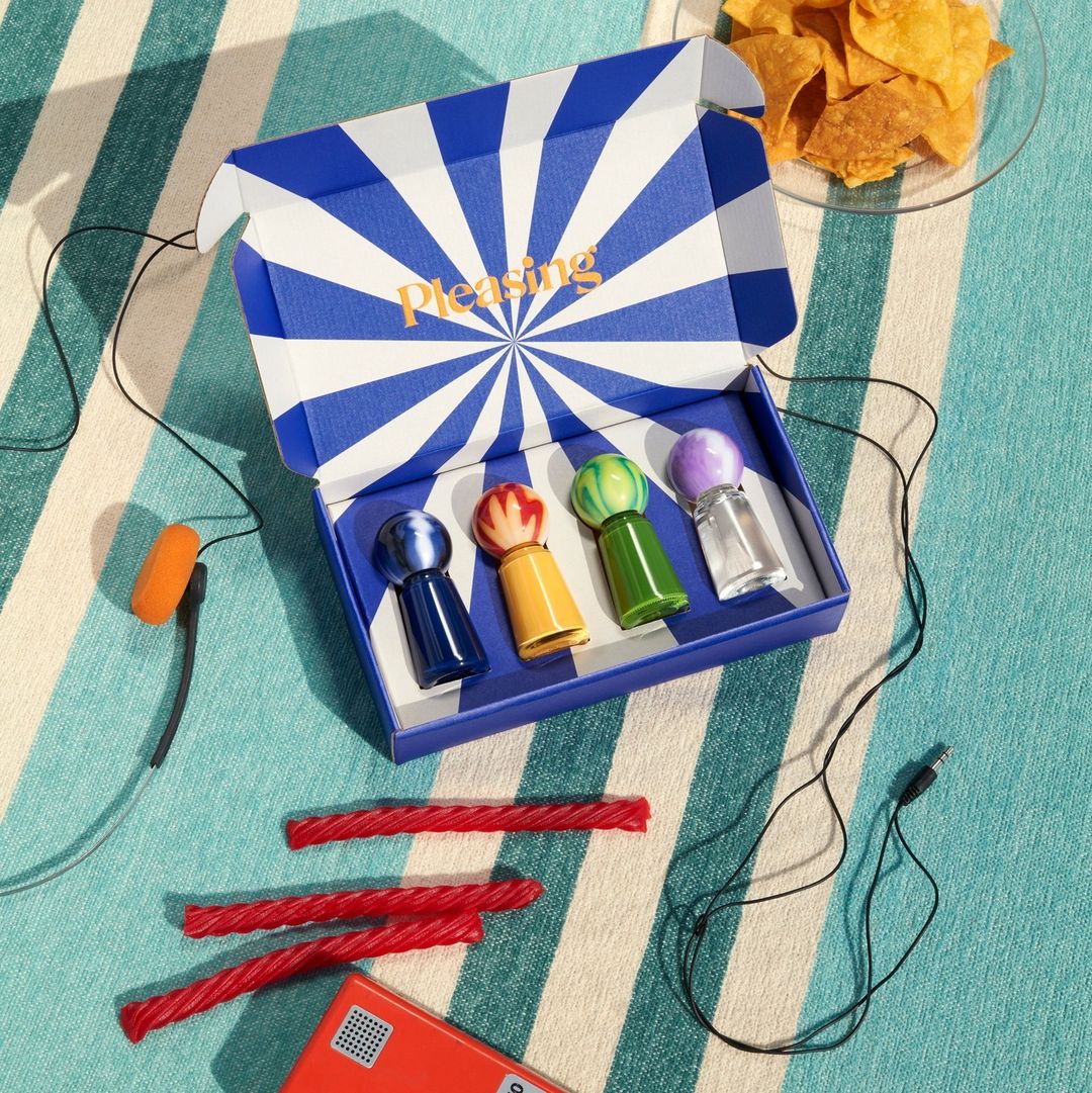 Close-up of 'Pleasing' beauty product box with colorful nail polish, chips, licorice, and retro tech on striped blue blanket.