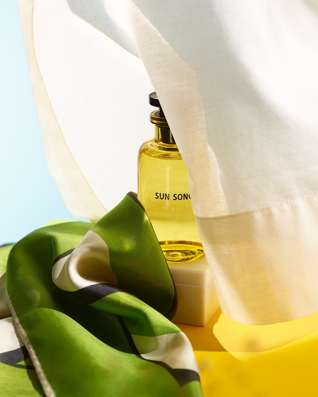 Still life shot of a yellow perfume bottle on a white platform, accented by a draped white cloth and a green and black scarf.