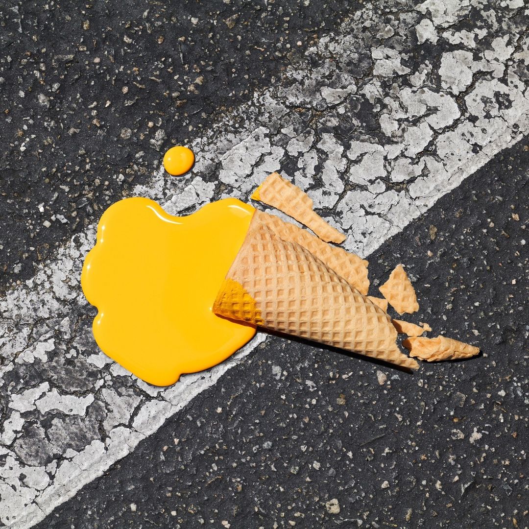 Overhead shot of melted yellow ice cream and broken waffle cone on asphalt surface with white line; food photography, still life.