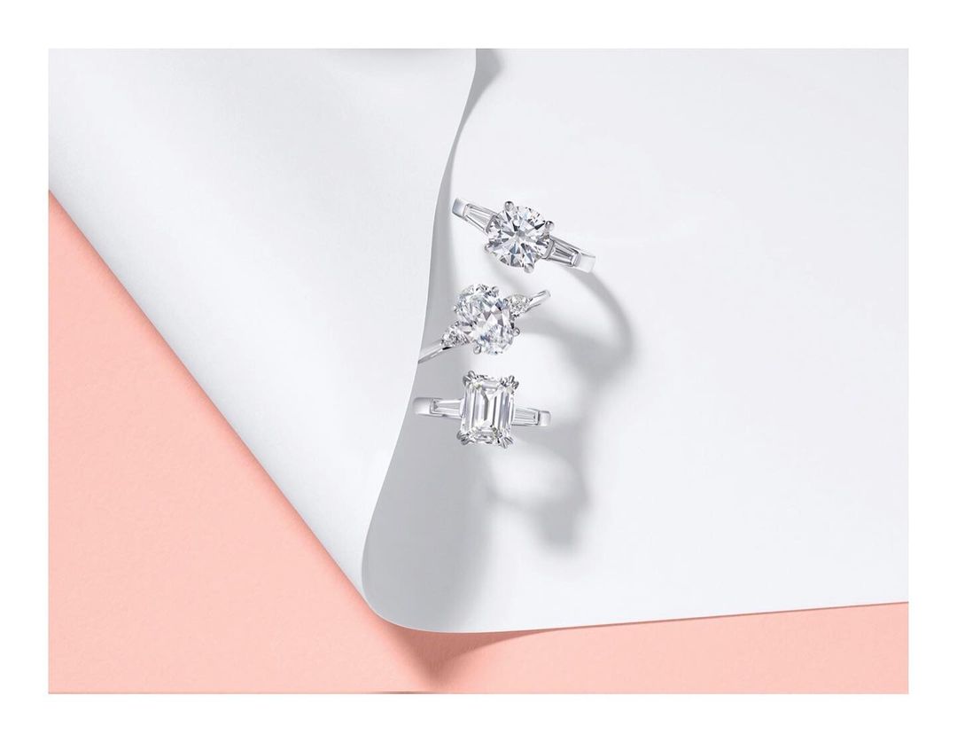 Three diamond engagement rings displayed on a white, folded paper with a light pink background, emphasizing their brilliance and design.