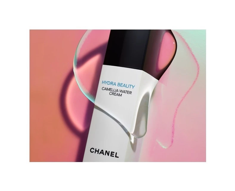 Close-up of a white bottle of Chanel Hydra Beauty Camellia Water Cream against a blurred pink and teal background with refracted light.