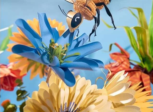 Close-up of paper art flowers in blue, yellow, and orange with a bee landing on the blue flower against a light blue background.