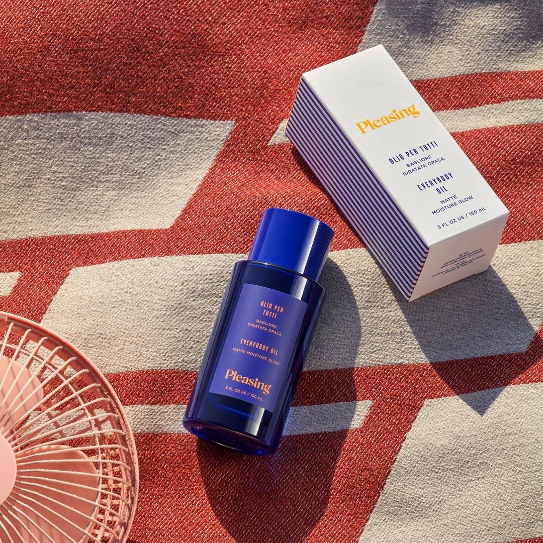 Pleasing brand's skincare products, including a blue bottle of body oil and a white box with blue stripes, are displayed on a patterned towel.
