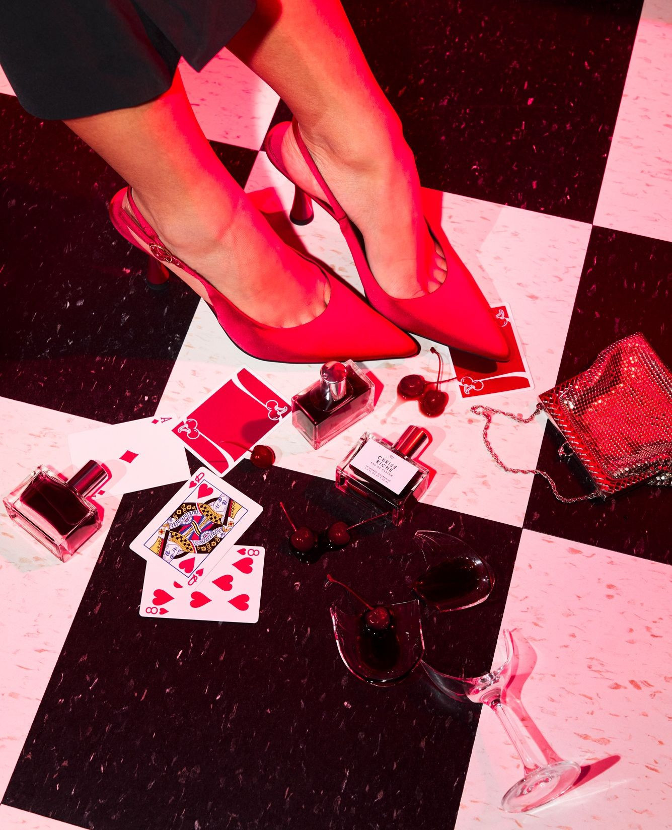 Woman's legs with red high heels and perfume bottles on a checkered floor, featuring playing cards and a broken wine glass.