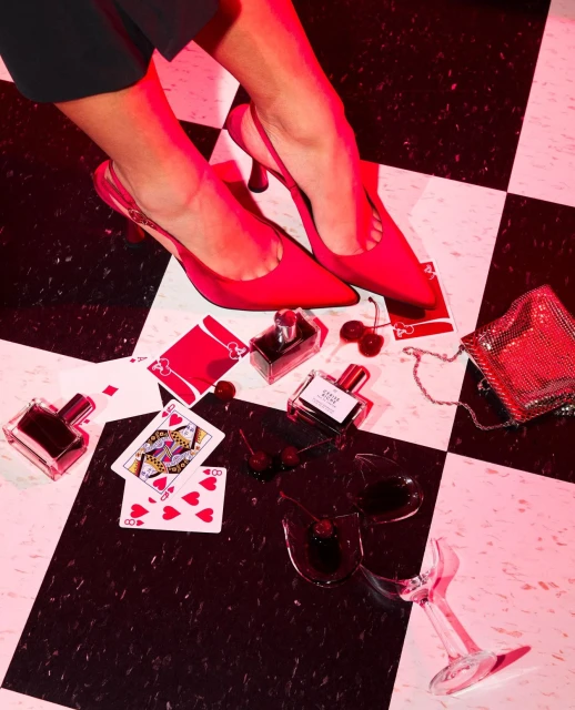 Woman's legs with red high heels and perfume bottles on a checkered floor, featuring playing cards and a broken wine glass.