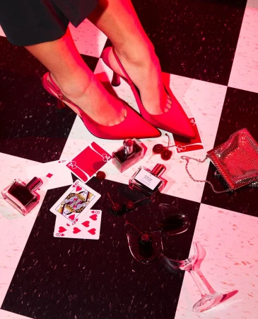 Woman's legs with red high heels and perfume bottles on a checkered floor, featuring playing cards and a broken wine glass.
