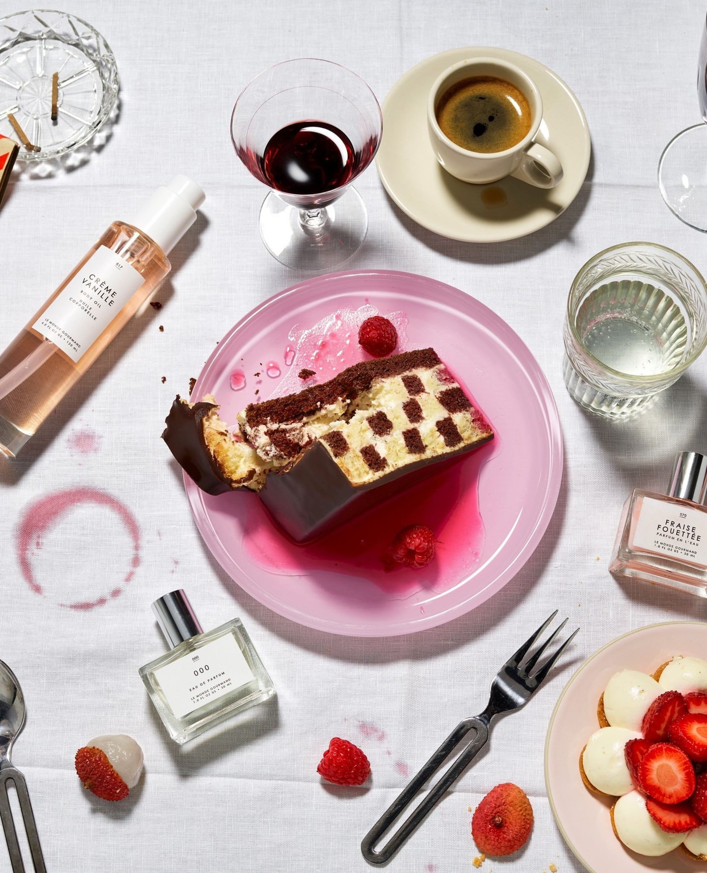 Overhead shot of a dessert plate featuring a slice of chocolate checkerboard cake, surrounded by perfume bottles, a wine glass, and coffee.