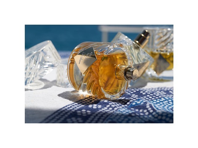 Luxury perfume bottles on a patterned cloth with hard shadows. Gold liquid is visible inside the transparent glass bottles.