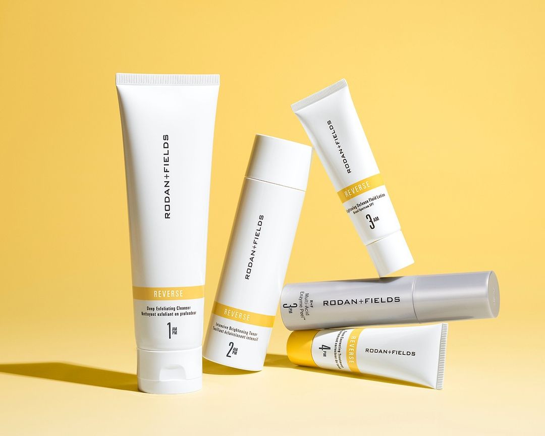 Several white RODAN+FIELDS REVERSE-branded skincare product tubes of different sizes are arranged in a still life against a yellow backdrop.