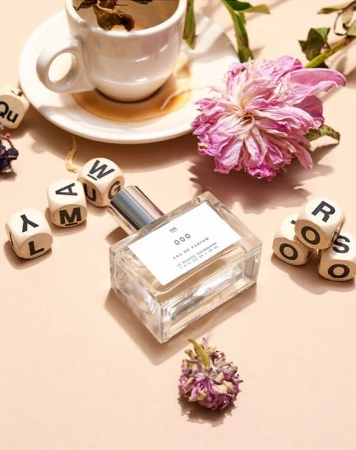 Still life of a clear glass perfume bottle on a beige surface with white ceramic cup, dried flowers, and lettered dice.