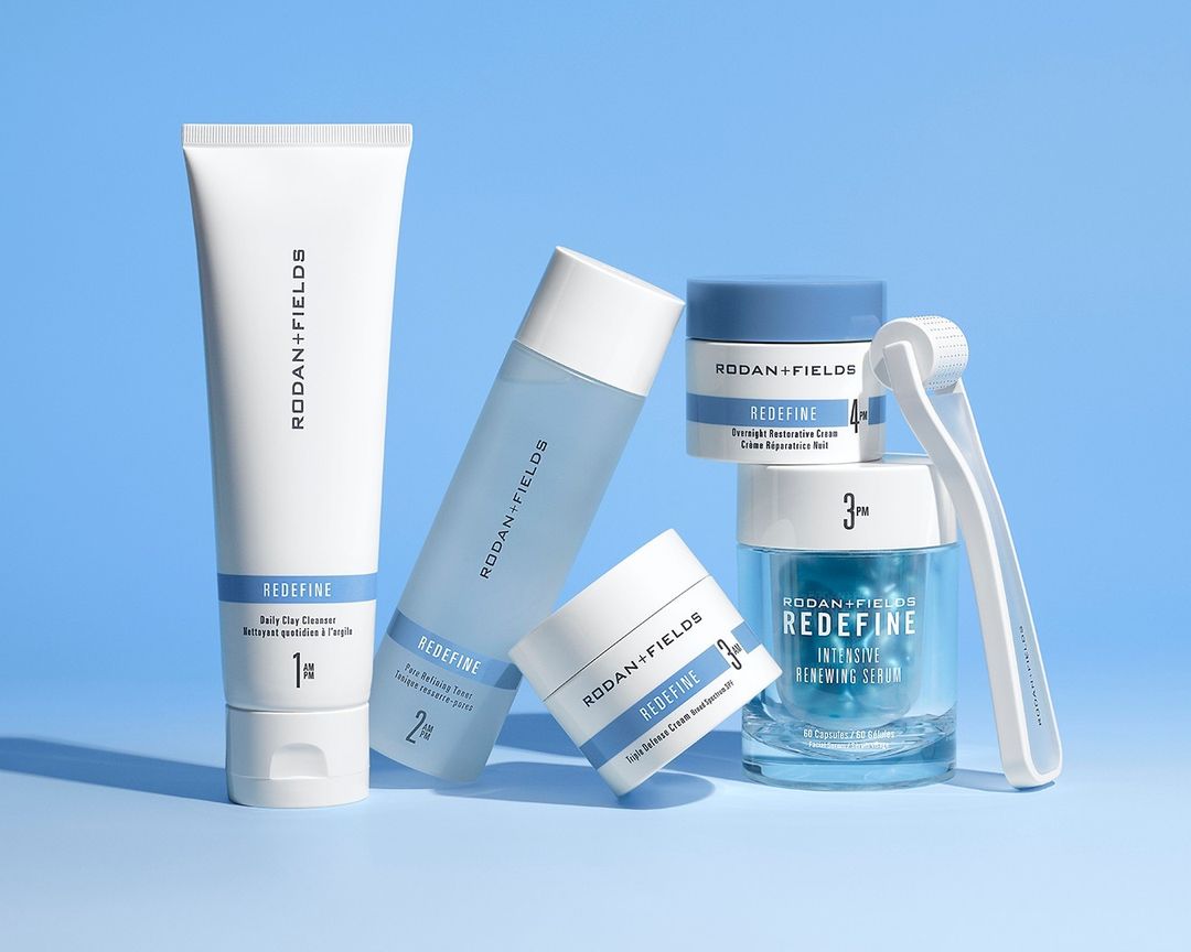 Rodan + Fields skincare product line featuring cleanser, toner, serum, and roller, arranged on a soft blue background in studio lighting.