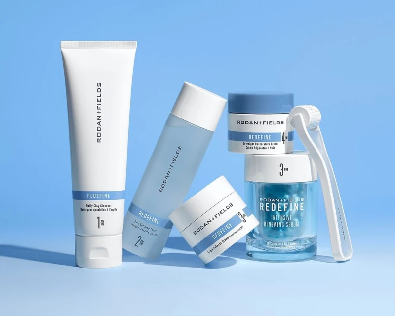 Rodan + Fields skincare product line featuring cleanser, toner, serum, and roller, arranged on a soft blue background in studio lighting.