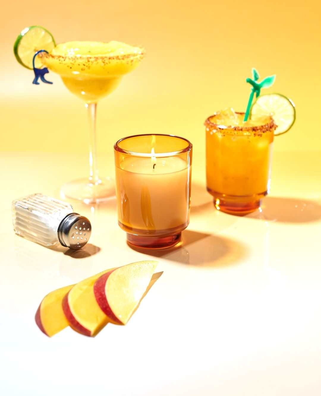 Two cocktails, one margarita in a stemmed glass and one in a rocks glass, with a candle, salt shaker and sliced fruit against a yellow background.
