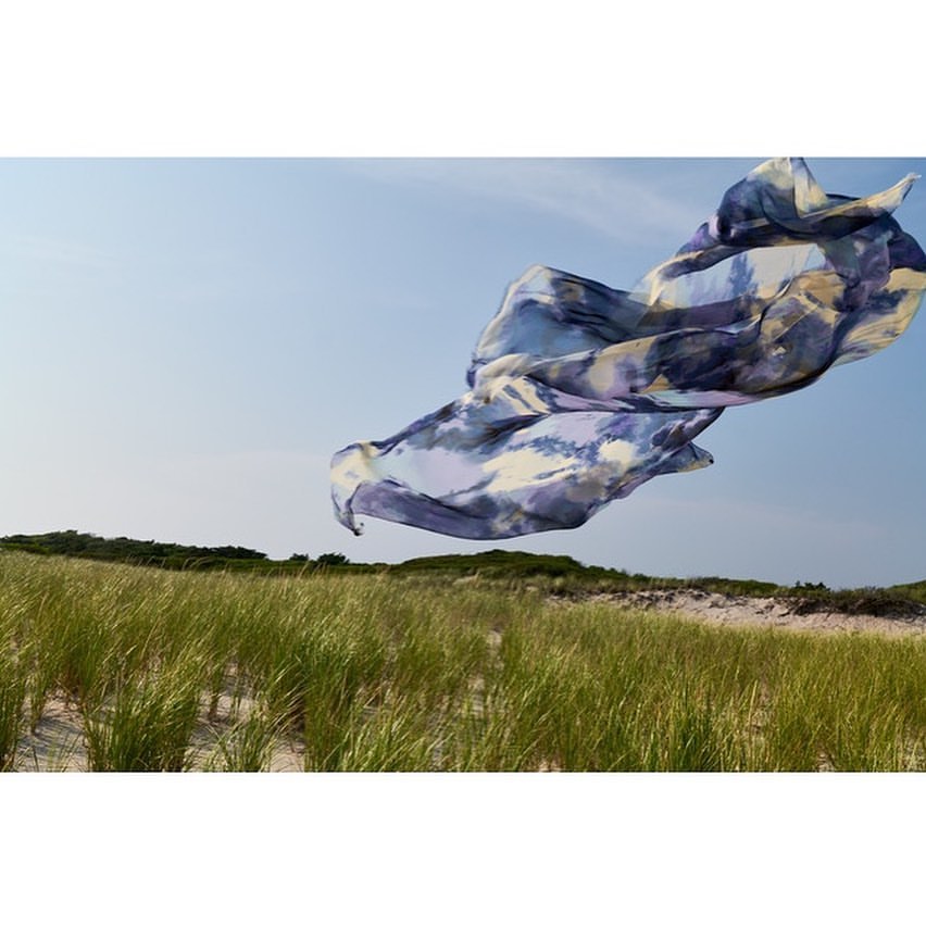 A fabric floats in the air over tall green grass growing on sand dunes underneath a blue sky. Landscape orientation, sunny day.