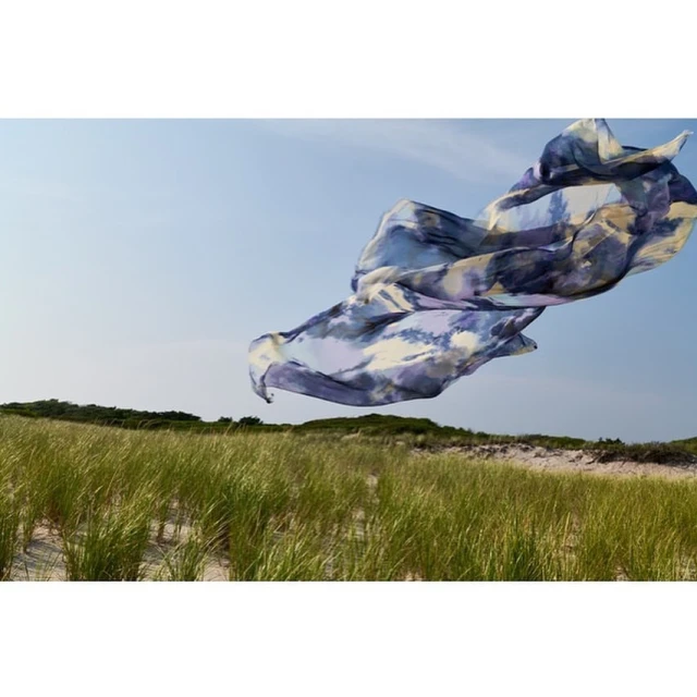 A fabric floats in the air over tall green grass growing on sand dunes underneath a blue sky. Landscape orientation, sunny day.