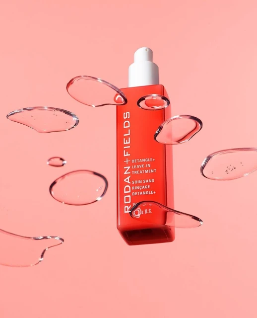 Red bottle of Rodan + Fields detangling leave-in hair treatment with floating water droplets on a light peach background.