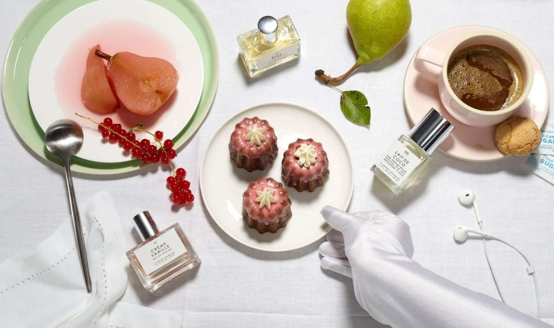 Still life composition with perfume bottles, desserts, fruit, and coffee creating a luxurious and sensory-rich arrangement on a white tablecloth.