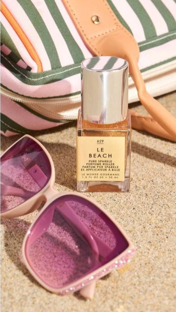 Close-up shot of a bottle of Le Beach perfume next to pink sunglasses and a striped tote bag on a sandy surface.