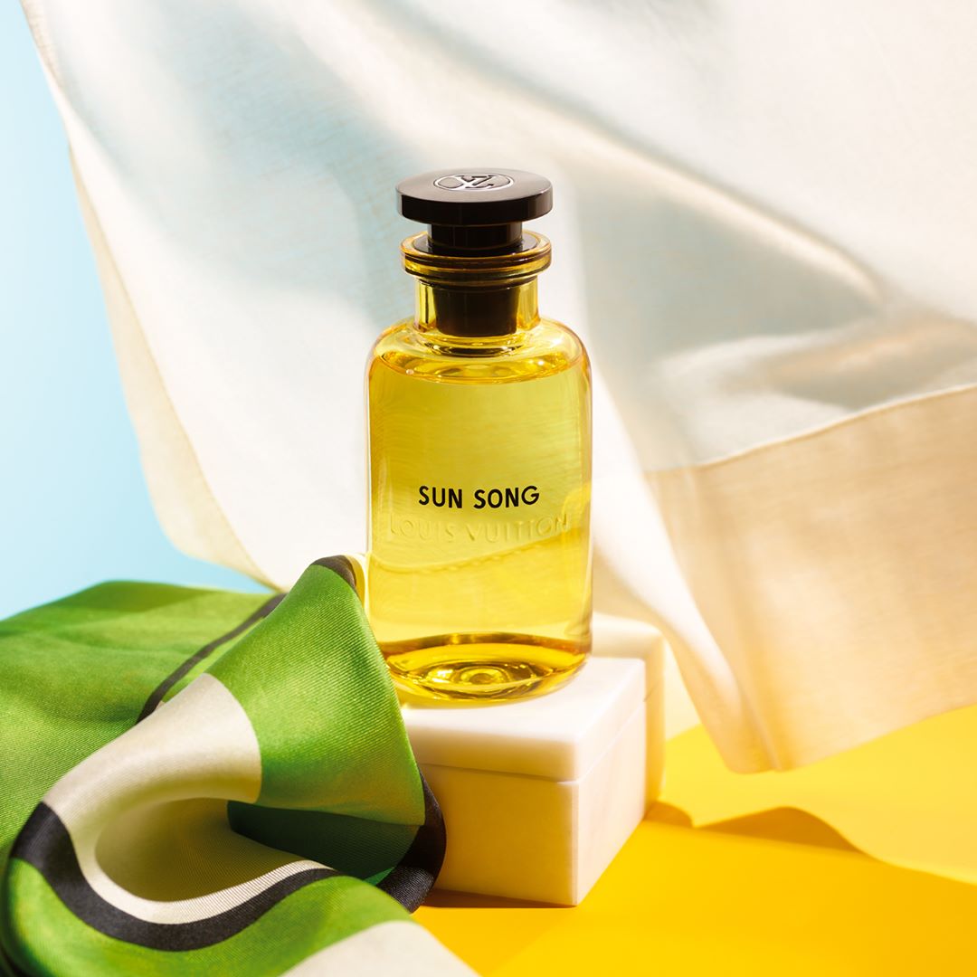 Still life shot of a yellow glass bottle of Louis Vuitton Sun Song perfume with a green silk scarf and minimalist backdrop, soft lighting.