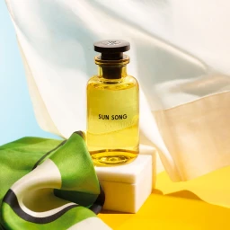 A glass bottle of Louis Vuitton Sun Song perfume stands upright on a white block with a green and white silk scarf in the foreground.