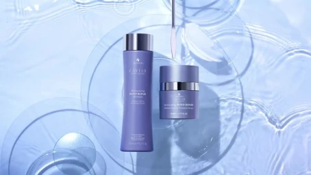 Two purple hair care product bottles and jars arranged on transparent glass discs over rippled blue water with a metal spatula.