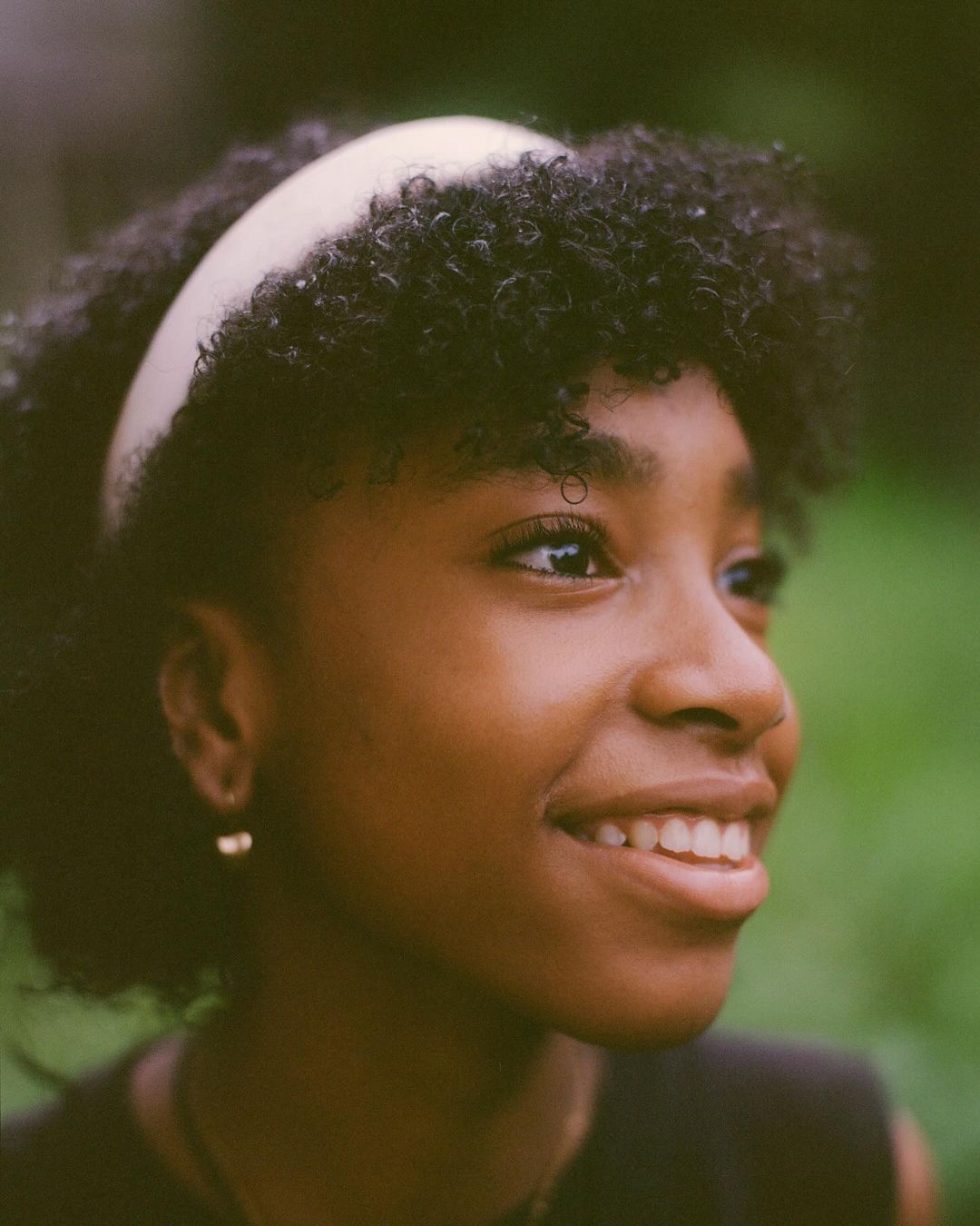 Close-up portrait of a young Black woman with natural hair and a headband, smiling in an outdoor setting with soft, filtered light.