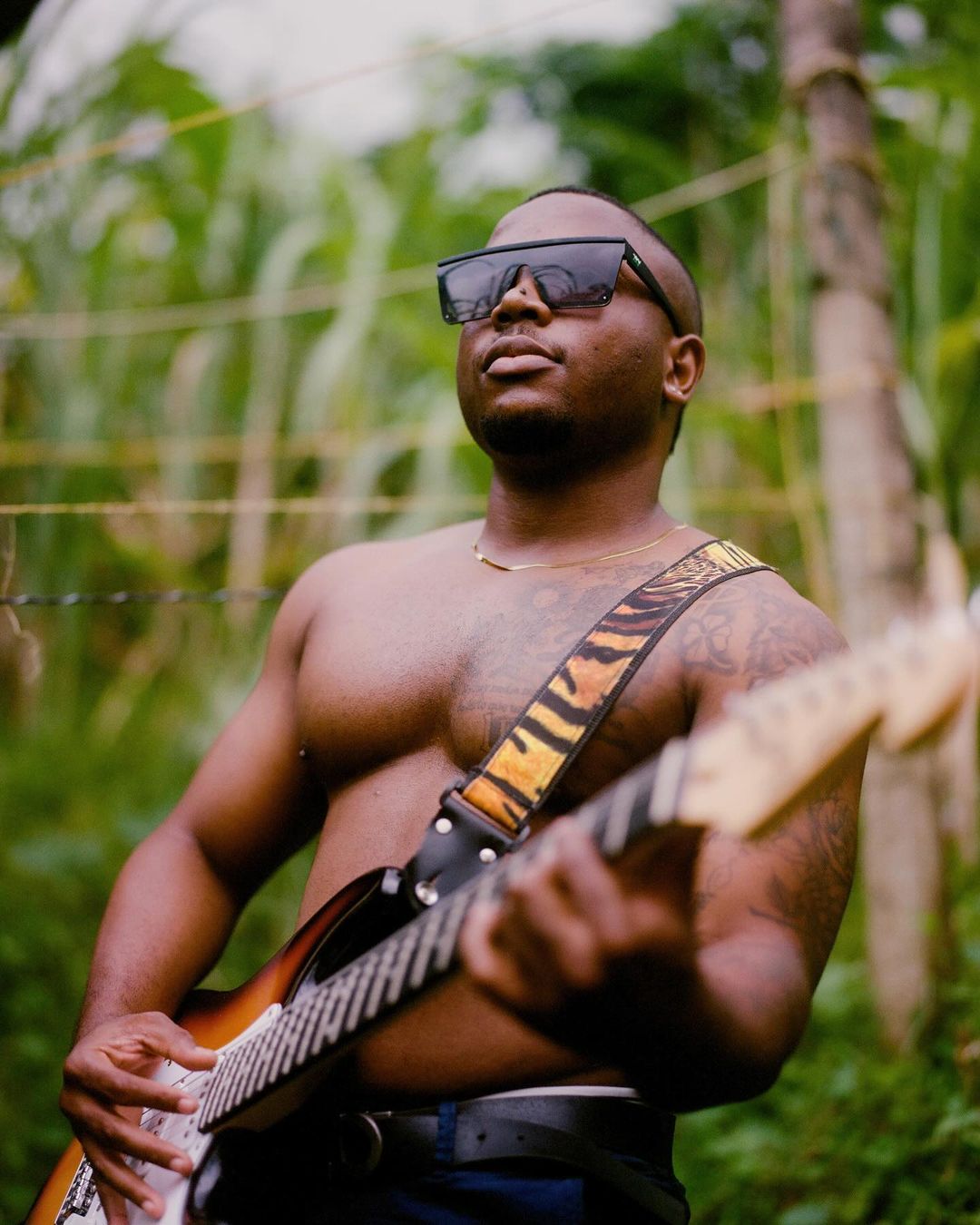 Shirtless Black man in sunglasses plays electric guitar in natural outdoor setting with greenery and a rustic fence creating a raw aesthetic.