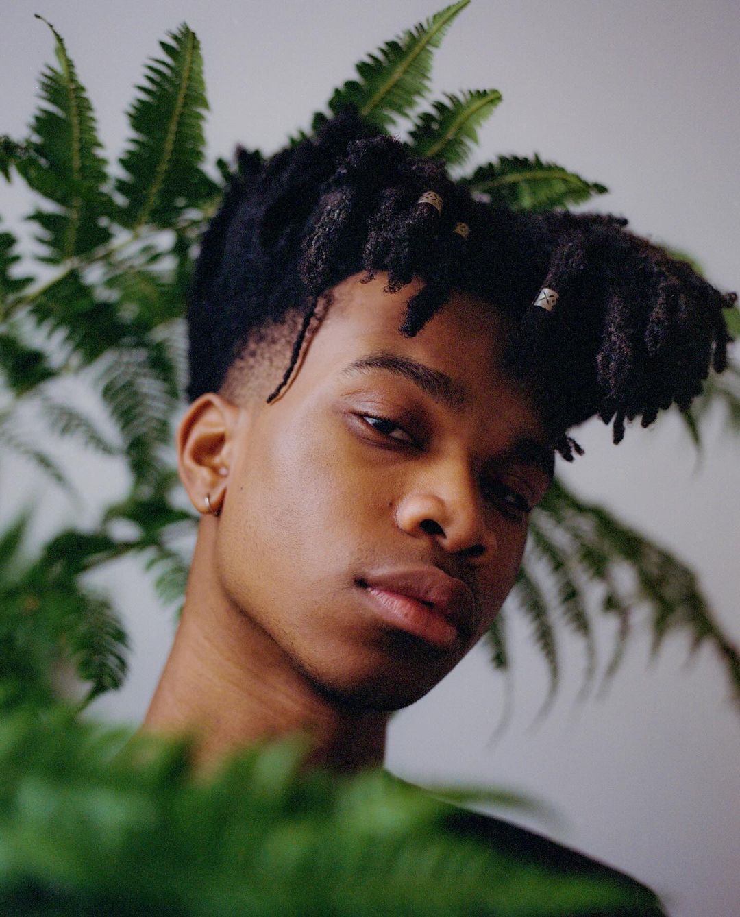 Portrait of a young Black man with dreadlocks partially surrounded by lush fern fronds in a bright, natural-light photo studio setting.