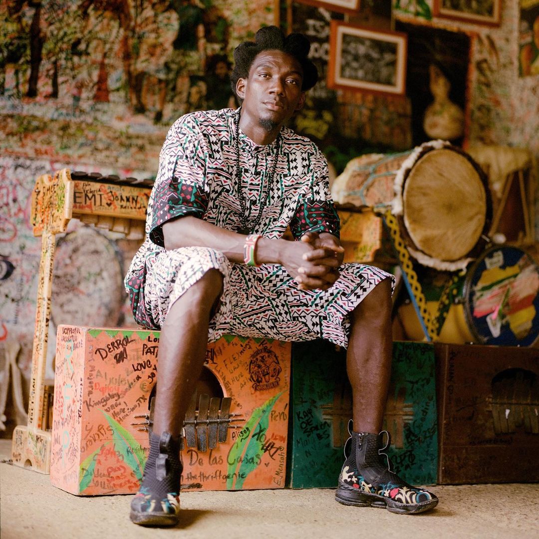 Adult male seated on painted boxes with musical instruments, wearing a patterned shirt and shorts in an indoor setting.