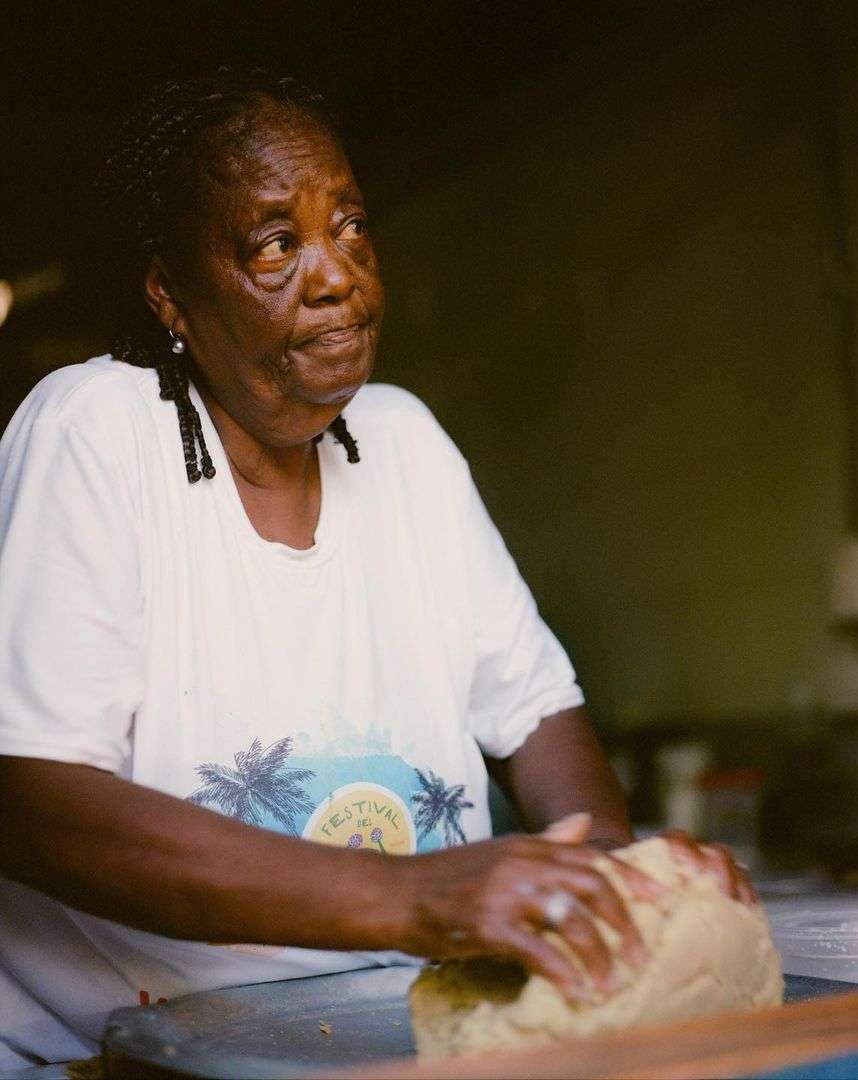 Elderly woman with dark skin and braided hair presses dough on a silver surface in an indoor setting with a vintage aesthetic.