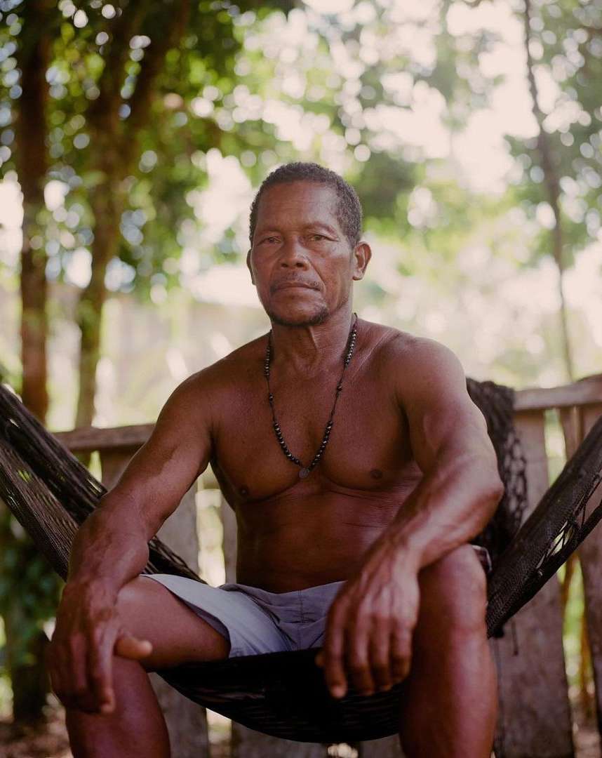 Portrait of an adult man sitting in a hammock outside surrounded by green trees, wearing shorts and a beaded necklace in natural light.