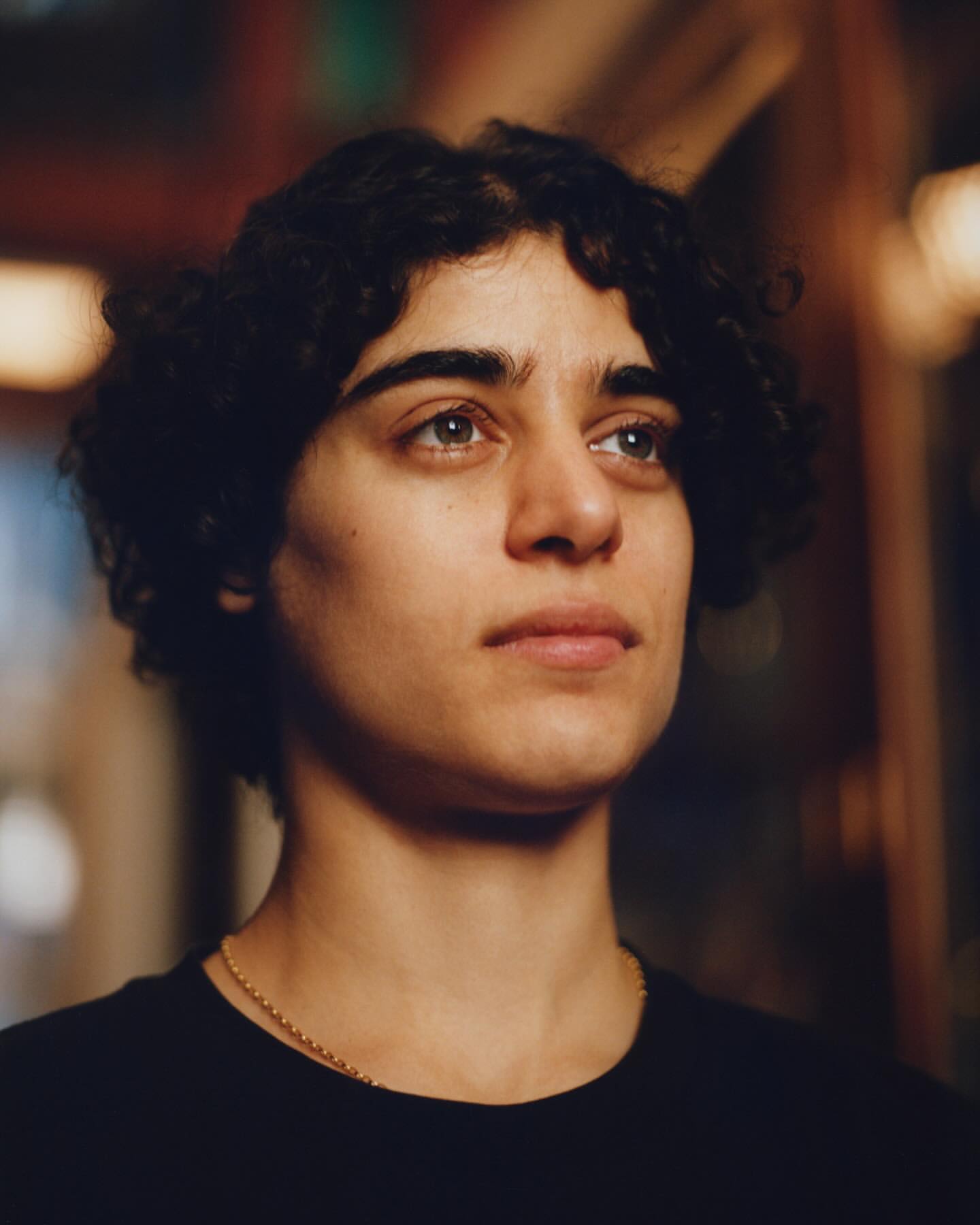 Close-up portrait of a young adult with curly hair looking up, wearing a black t-shirt and gold necklace, indoors with blurred background.