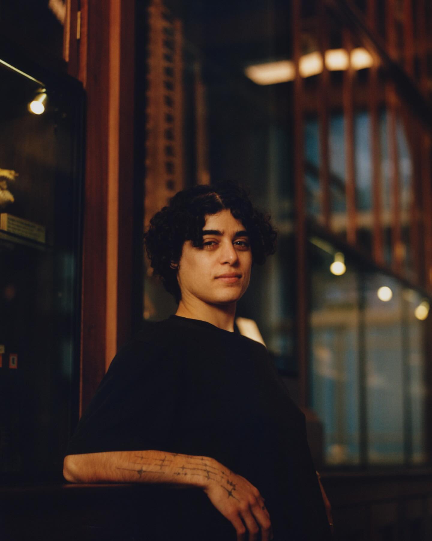 Portrait of a young adult with dark hair, wearing a black shirt, photographed indoors. The lighting is soft, and cabinets visible in background.