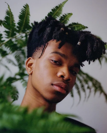 Close-up portrait of a Black man with dreadlocks framed by green fern leaves, photographed in London by Adïam Yemane.