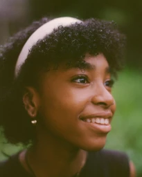 Portrait of a smiling young Black woman with a white headband outdoors in London, photographed by Adïam Yemane