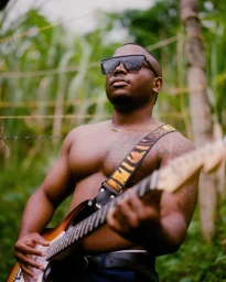 Black man wearing sunglasses and playing an electric guitar outdoors in a lush green setting, photographed by Adïam Yemane