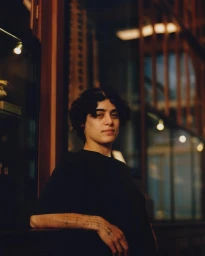 Portrait of a person with dark curly hair wearing a black t-shirt leaning on a counter indoors in London