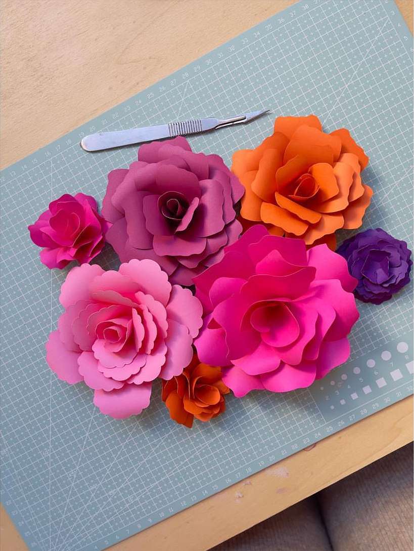 Crafted paper flowers in shades of pink, orange, and purple arranged with a craft scalpel on a self-healing cutting mat surface.
