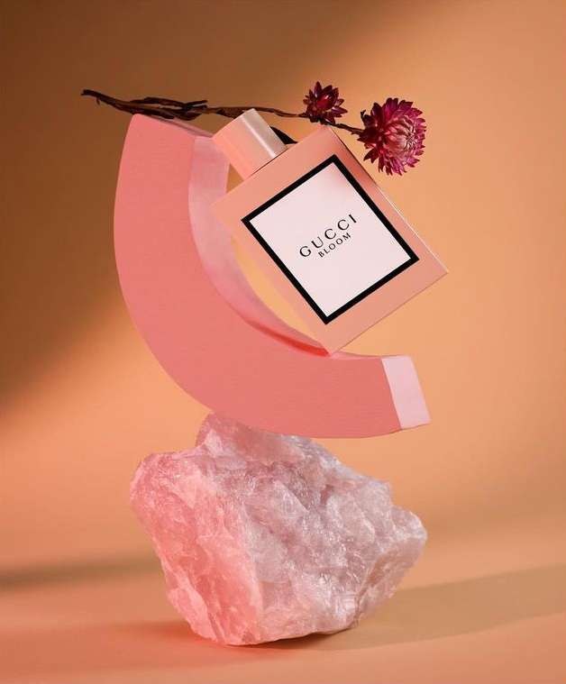 Gucci Bloom perfume bottle on a pink geometric stand and rock, adorned with dried flowers against an orange gradient background.