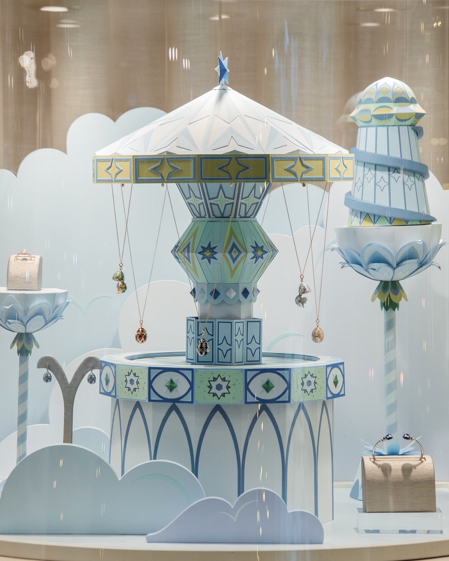 Carousel structure with hanging jewellery and decorative towers in shop display case against a light blue and white backdrop.