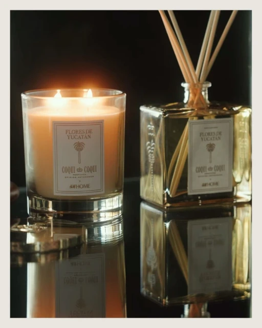 Illuminated Coqui Coqui Flores de Yucatan candle and fragrance diffuser set, set on a reflective black surface against a dark backdrop.