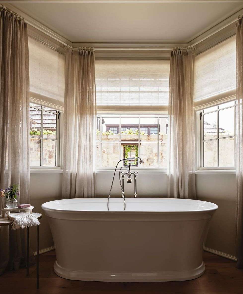 Freestanding white bathtub with chrome faucet in a neutral-toned bathroom with natural light from large windows and sheer curtains.
