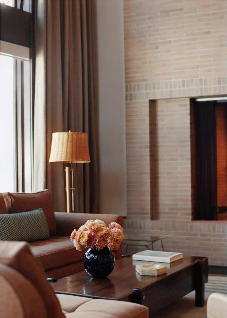 Living room interior with brown sofas, a coffee table with flowers, a brick fireplace, and window light filtered by brown curtains.
