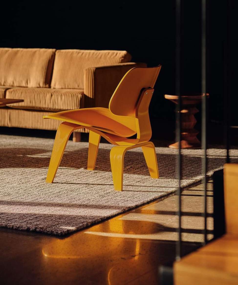 Interior view featuring a modern yellow plywood chair, a tan corduroy couch, a round side table, and a light gray rug on a brown floor.