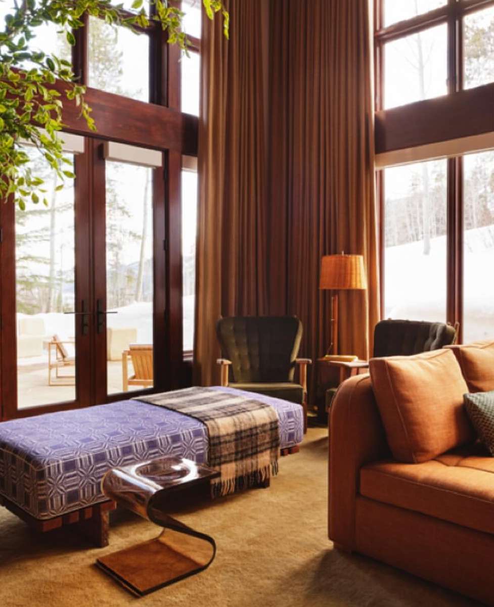 Interior view of a living room with an orange sofa, purple patterned daybed, and large windows looking out onto a snowy landscape.
