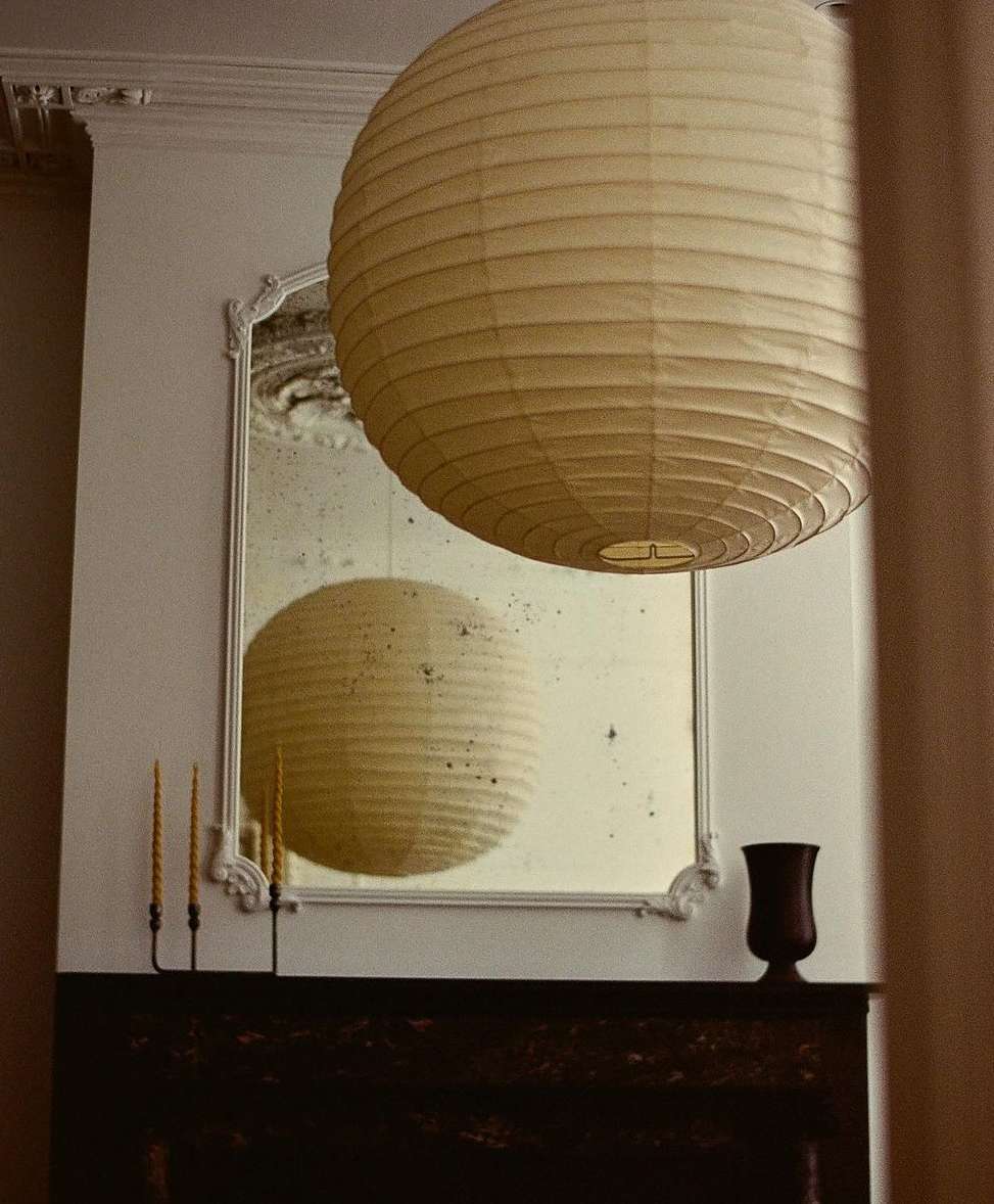 Interior with cream paper lantern, antique mirror reflecting the lantern, black fireplace mantel, cream walls, and tapered yellow candles.